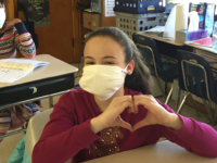 A student at Adams Elementary School wears their new face mask and holds their hands in the shape of a heart to express their gratitude.
